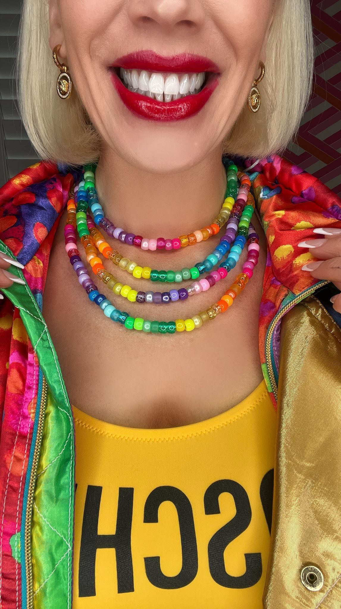 Blonde lady in yellow vest and colourful jacket wears stacked beaded rainbow necklaces.